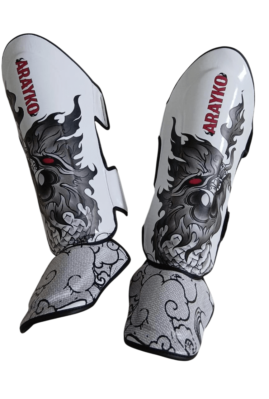 Pair of professional white Muay Thai shin guards showcasing a gray Thai dragon with red eyes, ARAYKO branding, and decorative cloud-patterned foot protection